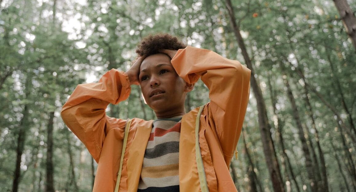 Teen holding head in thinking pose, wearing yellow raincoat, surrounded by forest greenery.